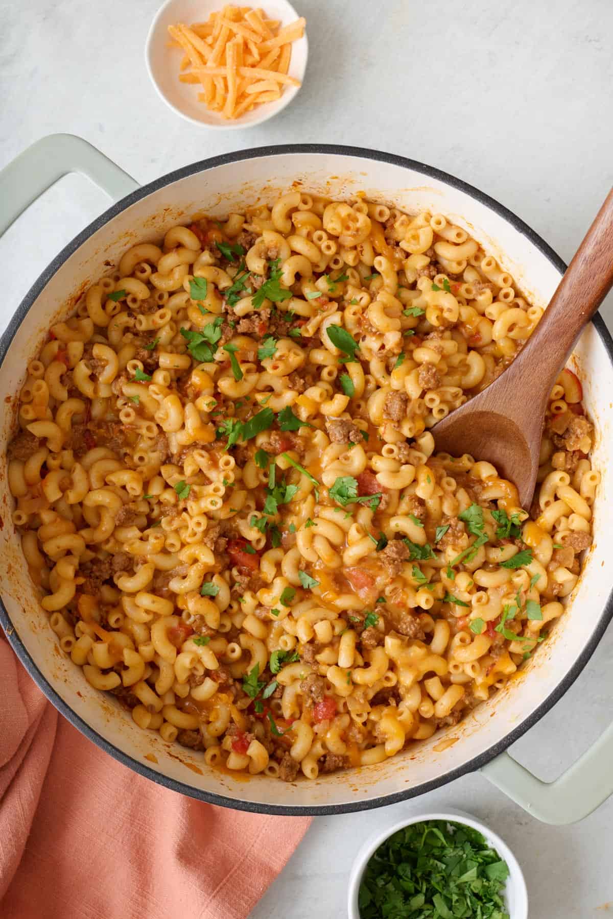 Dutch oven of taco pasta with a wooden spoon with fresh cilantro and shredded cheese nearby.